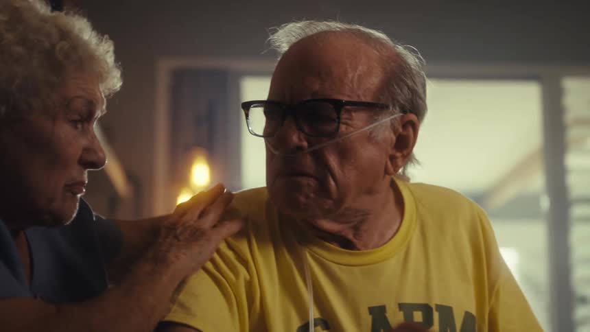An elderly woman with gray hair places her hand on the shoulder of an elderly man wearing a yellow tshirt and an oxygen tube in his nose as they sit together