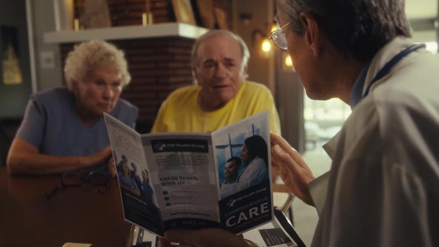 A doctor wearing a white lab coat a stethoscope around his neck and glasses holds a pamphlet showing it to an elderly couple seated across the table as he explains it to them