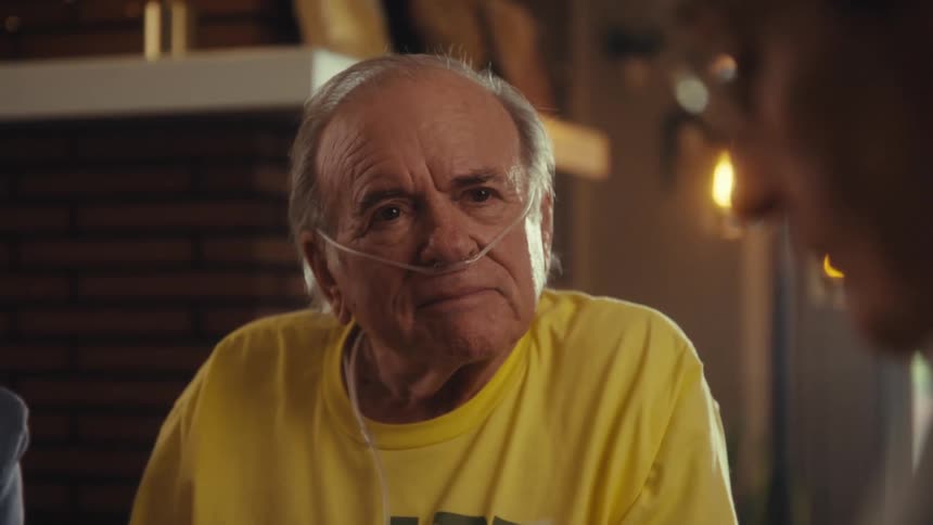 An elderly man wearing a breathing aid listens to another man talking as the other man pulls out a pamphlet