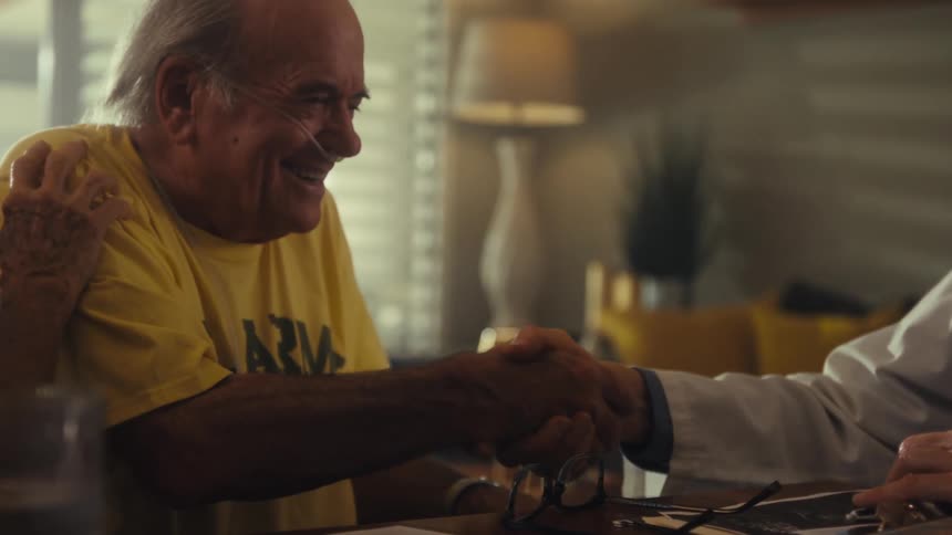 A doctor wearing a lab coat with a stethoscope over his shoulder talks to an elderly couple he shakes the elderly mans hand causing excitement as the woman begins speaking the elderly man is wearing a nasal cannula