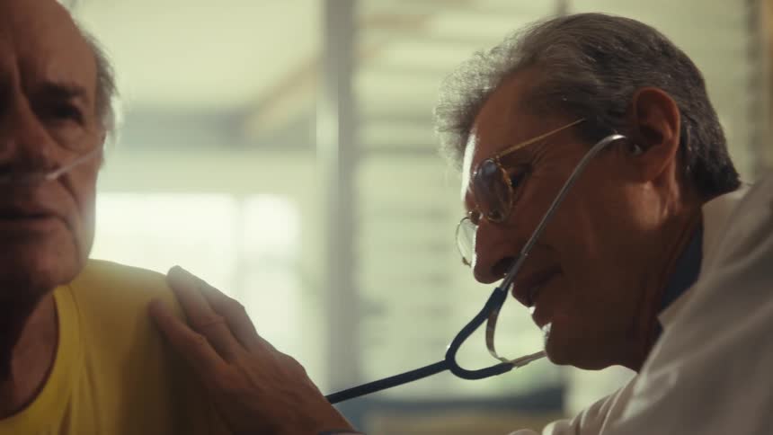 A doctor using a stethoscope to check an elderly mans heartbeat with his wife beside him offering support