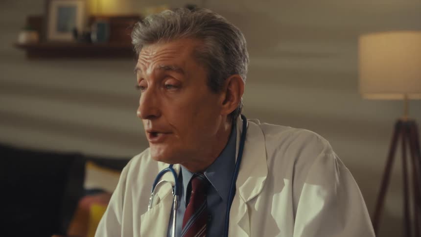 A medical professional wearing a white coat a dark tie and a stethoscope holds a black tablet in his left hand looks from side to side and then talks while making a serious face