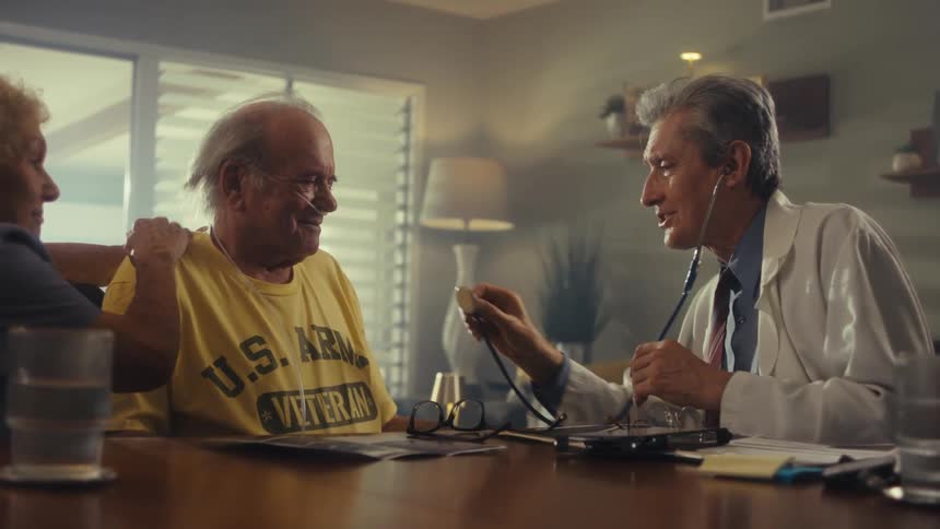 A medical professional wearing a stethoscope and a white coat is examining a man who is sitting at a table the man is wearing a yellow shirt with us army veteran written on it and has what appears to be a nasal cannula the medical professional uses