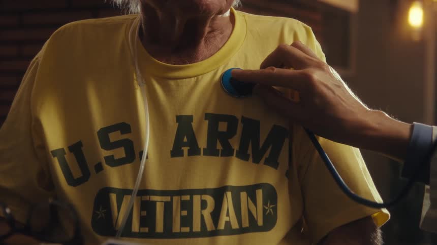 A doctor uses a stethoscope to check the heartbeat of an elderly man wearing a yellow us army veteran shirt and a nasal cannula