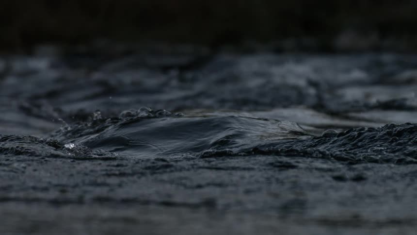 A slowmotion closeup video captures the hypnotic and moody movement of dark water showing smooth ripples and eddies forming on its surface