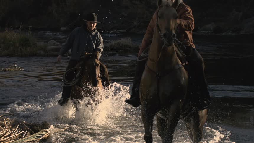 Two cowboys ride horses through a shallow river at dusk