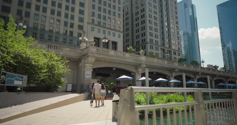 Couple walks with a dog on a riverside walkway surrounded by tall modern buildings