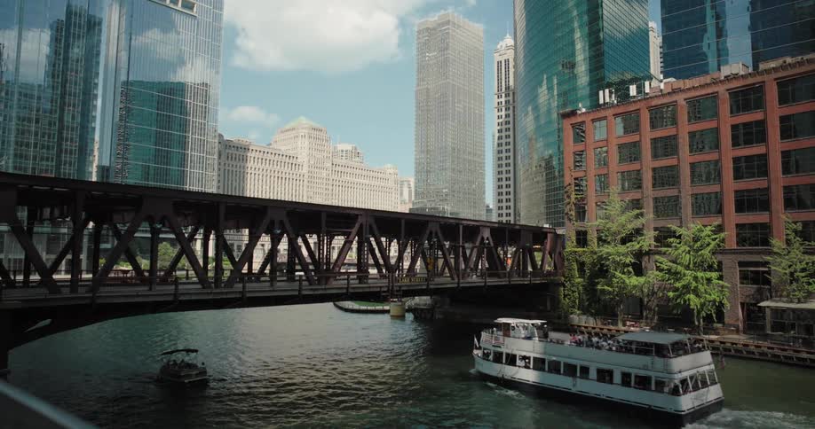 A boat moves along a canal and passes under a bridge with city buildings highrises and skyscrapers lining both sides at midday