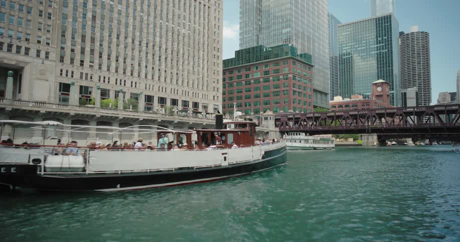 A large tour boat with tourists on a river beside skyscrapers and a bridge