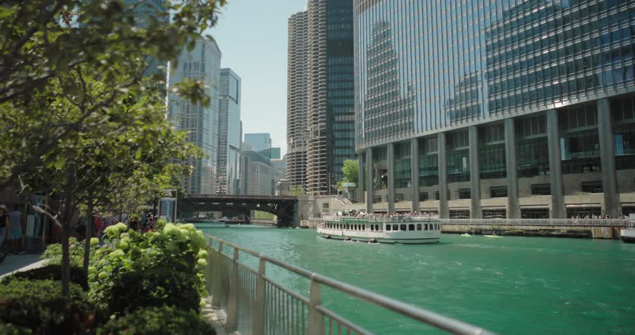 A view of the chicago river is shown with a tour boat sailing down the tealcolored water the river is surrounded by modern buildings and a walking path with lush greenery and a metal fence a bridge can also be seen spanning the river in the background