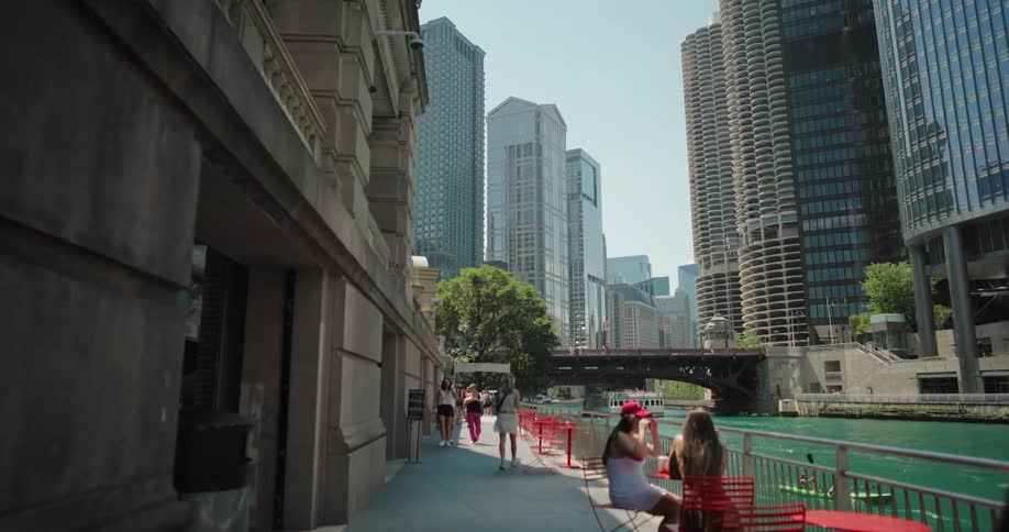 People are seen sitting at red tables and walking on a sidewalk next to a river in a large city with many tall buildings