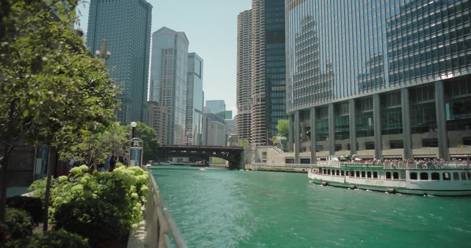 A view of the chicago river is shown with a boat sailing down the tealcolored water the river is surrounded by modern buildings and a walking path with lush greenery and a metal fence a bridge can also be seen spanning the river in the background