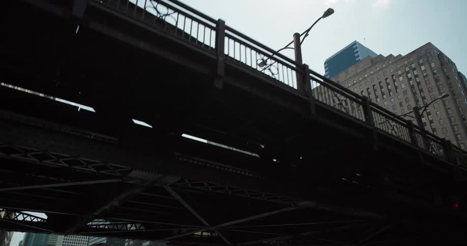 A camera travels underneath a large dark metal bridge then emerges to reveal a view of tall city buildings under a bright sky with a river and a boat full of people visible below