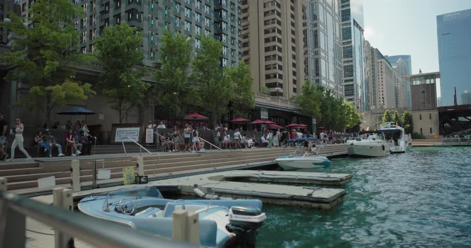 People walk and rest by the riverside with docked boats and tall buildings in the background