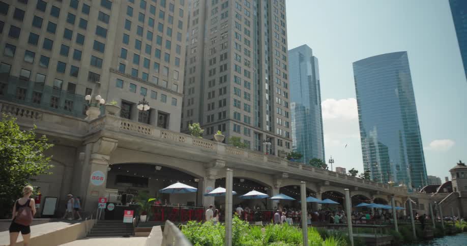 A bustling riverside promenade with outdoor dining and modern buildings rises under a clear sky
