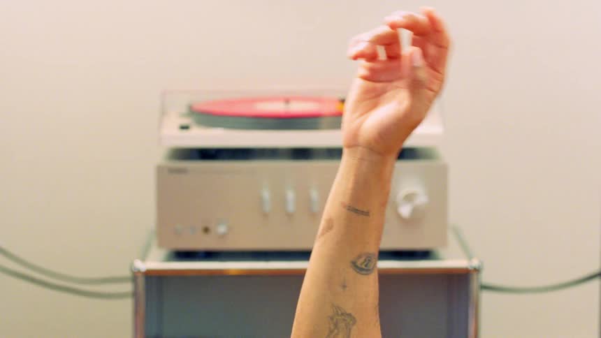 A turntable with a red record spinning on it is stacked on top of a silver amplifier with a persons tattooed arm and hand rising up from the bottom of the frame and waving its fingers back and forth