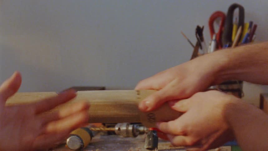 Two people in a carpentry workshop use sandpaper to smooth the surface of a piece of wood at midday
