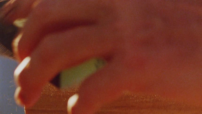 A persons hand shaves a thin curl from the edge of a wooden plank with a sharp tool