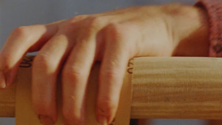 A close view of a couple persons hands shows them holding a piece of sandpaper and rubbing it back and forth on a piece of wood