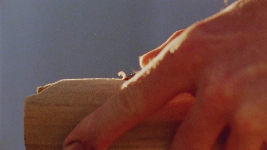 A closeup shot shows a persons hands holding a piece of lightcolored wood another hand is using a tool to scrape a thin layer of the wood off the surface which sends a small amount of sawdust flying into the air