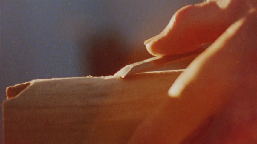 A closeup shot shows a persons hands carving or shaping a piece of wood a small lightcolored curl of wood shavings is visible indicating that the tool is effectively removing material from the block