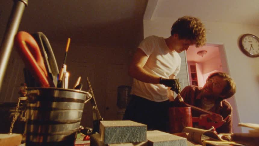 A man paints a piece of wood while another man watches beside him and both laugh during the process indoors