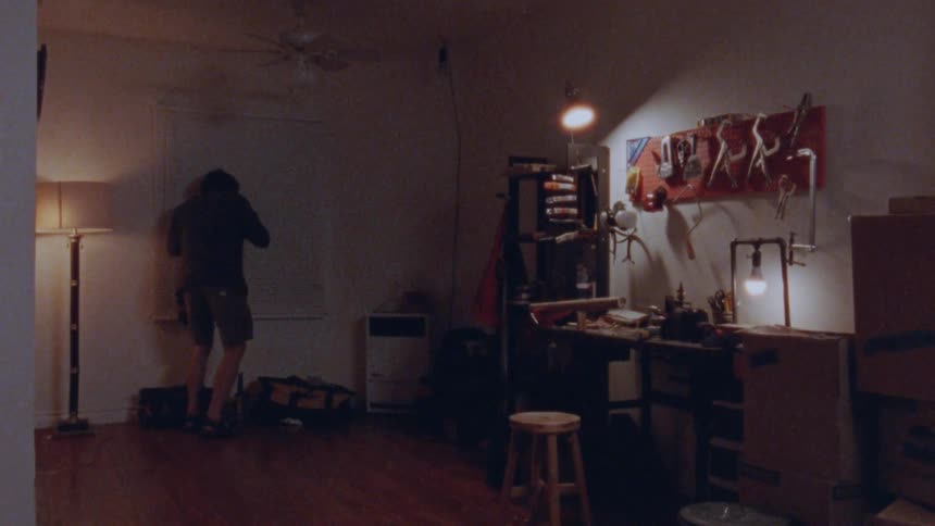 A person wearing a dark jacket and light shorts is sitting at a workbench with a variety of tools hanging on a red wall and then stands up to walk to the other side of the room talking nervously on the phone