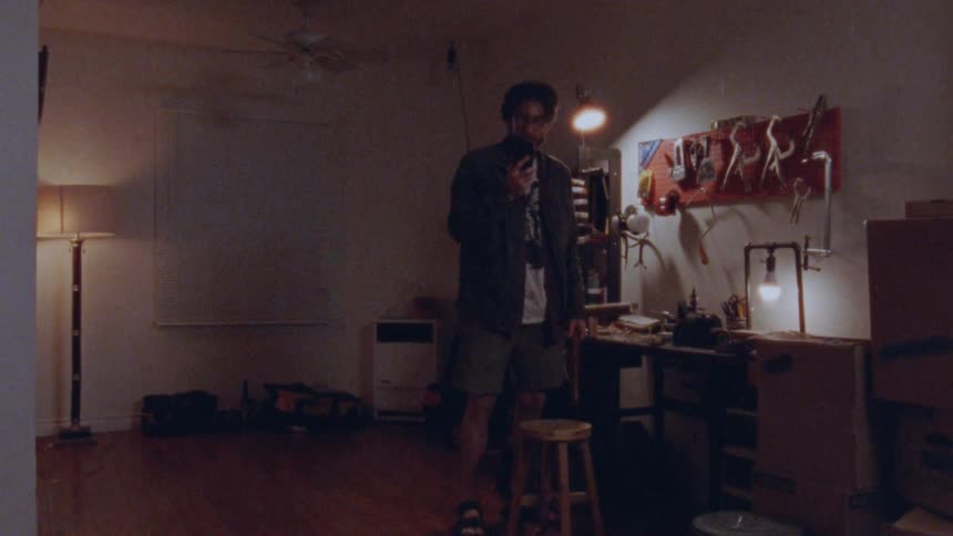 A man stands in a dimly lit room with a workbench and tools as he holds a smartphone to his ear and walks back and forth