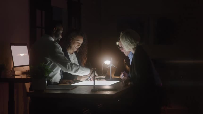 Three people gathered around a table lit by a desk lamp reviewing documents in an office