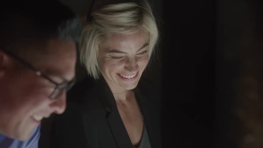 The video shows a man and a woman sitting close together smiling and laughing in a dark room the light from a screen below them illuminates their faces highlighting a sense of shared joy or camaraderie