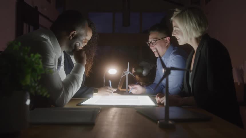 Four people in an office having a meeting at night sitting at a table with a light box with windmill models on the table