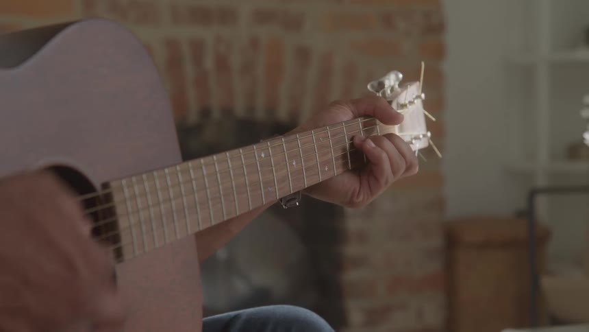 A person strums an acoustic guitar by a brick fireplace