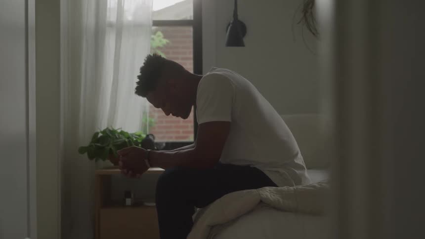 A man sits on the edge of a bed his head lowered in a sad despondent look