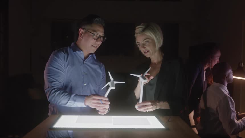 Two people examine wind turbine models on a lit table
