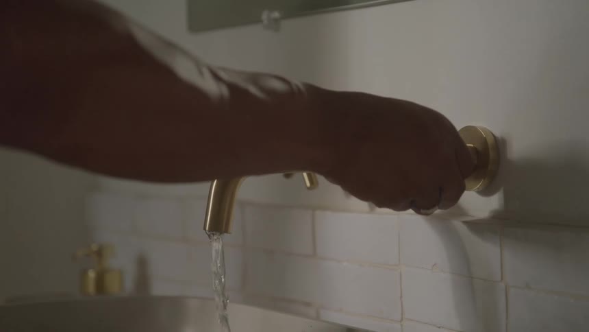 A close view shows a persons hand turning a gold faucet knob as water begins to flow from the faucet and a gold liquid soap dispenser sits on the counter below