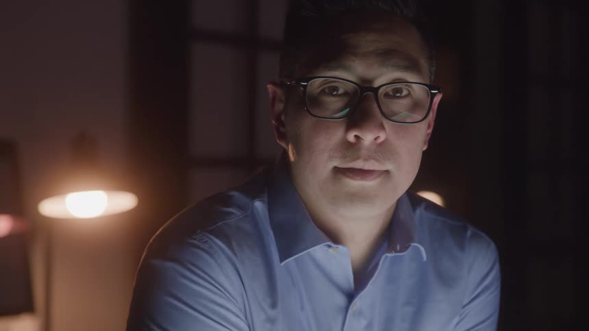 Man with glasses and a serious expression looks forward in a room lit by lamps