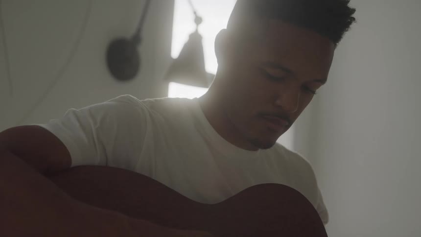 A black man in a white shirt plays a guitar and looks off to the distance