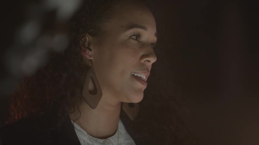 Close up portrait of a young black woman conversing with coworkers in a room at night