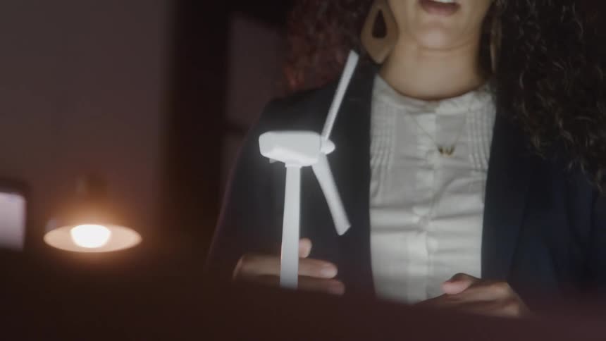 A business professional wearing is seen from the chest up with her long dark curly hair visible as she gestures with her hands toward a small white wind turbine