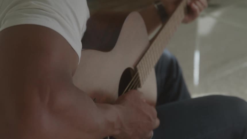 A person with a fade haircut and a white shirt is shown playing an acoustic guitar the video focuses on their hands and the guitar as they strum the strings