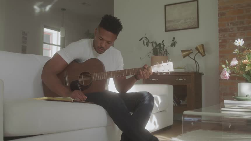 A musician is songwriting while playing guitar on a white couch