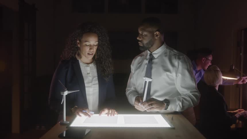 The video shows a group of business professionals in a dimly lit office setting working late a man and a woman in the foreground are intently looking at a litup light box on a tablethey appear to be discussing a project with the woman gesturing at