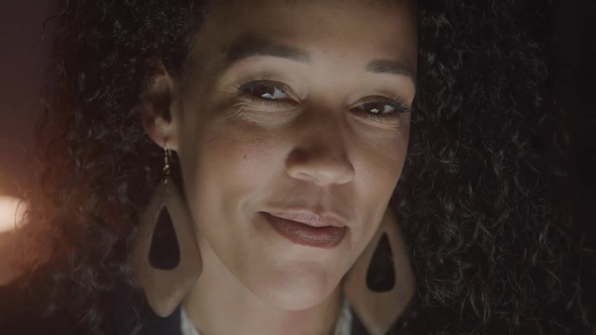 A woman with dark curly hair and long teardrop shaped earrings looks down then slowly raises her eyes and smiles faintly at the camera