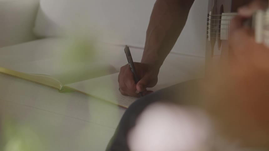 A person is seen writing in a notebook with a pen then switches to playing an acoustic guitar