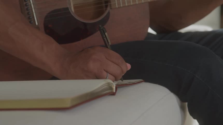 Man sitting on couch with a guitar writing lyrics in a journal