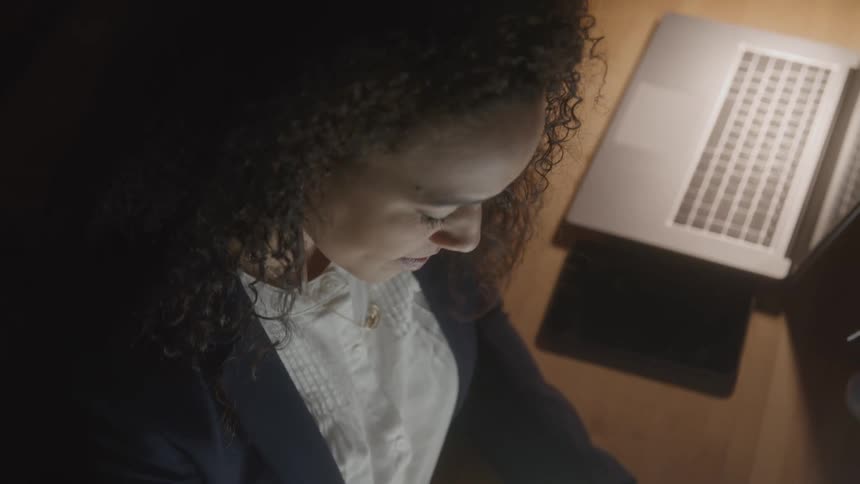 A woman with curly hair wearing a white shirt and dark jacket looks down intently illuminated by a light source offscreen with a laptop and a dark tablet visible on a wooden surface beside her