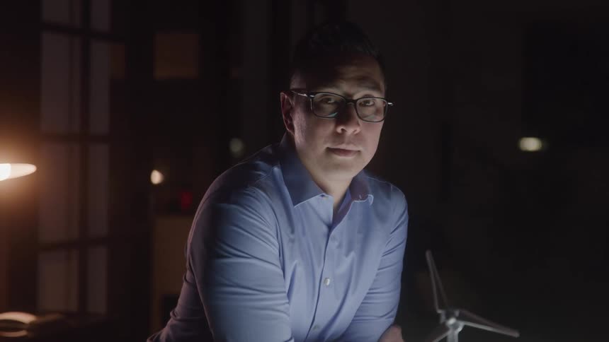 Man wearing glasses in a dark room looks forward with a serious expression