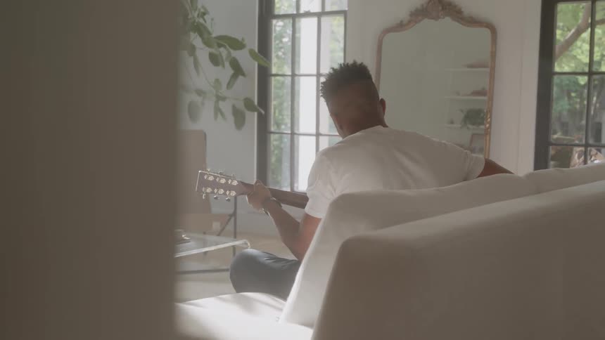 A young man dressed in white sitting on a white couch in a mostly white room playing guitar