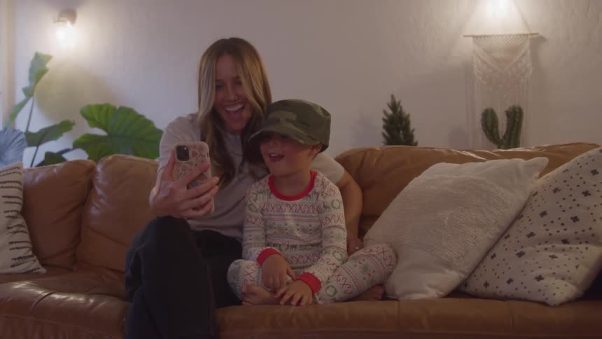 A mother and her son sit on a couch the boy wearing a comfortable hat both smiling as they look at a phone during a video call