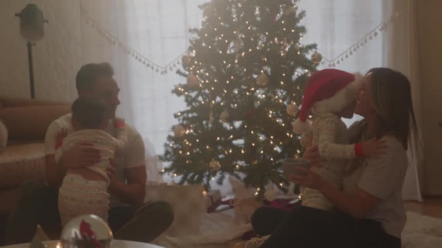 A family of fourtwo toddler boys a father and a mothersits on the floor in front of a christmas tree as each boy excitedly hugs one parent after the other